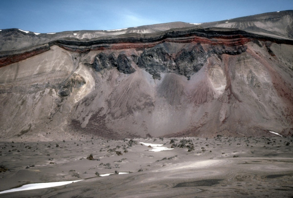 View of the cross section of Half Cone, a postcaldera vent now exposed against the north wall of Aniakchak caldera. Within this cliff face are recorded several episodes of lava flows (dark rock outcrops), plinian eruption (lighter colored deposits at left), and spatterfed-flow accumulation (reddish layers).  Photograph by R. McGimsey, U.S. Geological Survey, July 1, 1992.