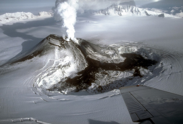 Steam rising from the intracaldera cinder cone at Veniaminof volcano in the waning stages of the 1983 to 1984 eruption. Cooling lava flows fill a pit about 2.3 1.0 km (1.4 0.6 mi) that has been melted in the summit ice cap. Aerial view looking northeast. Photograph by M.E. Yount, U.S. Geological Survey, January 23, 1984.