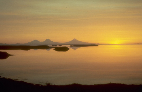 Sunset view to the southwest silhouetting Roundtop, Isanotski, and Shishaldin volcanoes on Unimak Island in the eastern Aleutian Islands. Photograph by J. Davies, Geophysical Institute, University of Alaska Fairbanks, August, 1983.