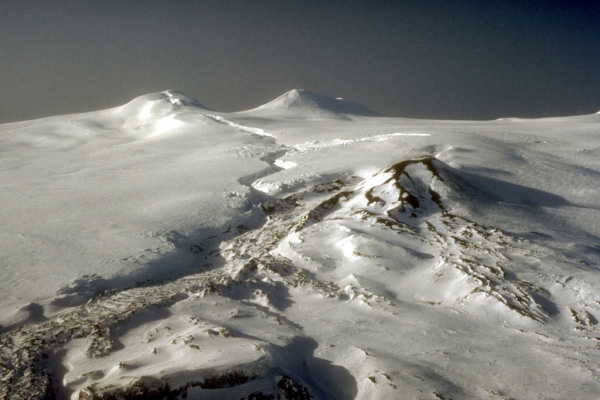 Summit area of Westdahl volcano located on the southwest part of Unimak Island in the eastern Aleutian Islands. The topographic peaks in the distance are Westdahl Peak (left) and Faris Peak (right). The cinder cone in middle ground marks the principal vent for the 1991 to 1992 eruption. Note the snow-mantled lava flow emanating from cinder cone. The sinuous fissure cutting the ice cap formed in the opening phases of the eruption and was the location of spectacular lava fountaining. Photograph by Fred Zeillemaker, U.S. Fish and Wildlife Service, February 2, 1993.