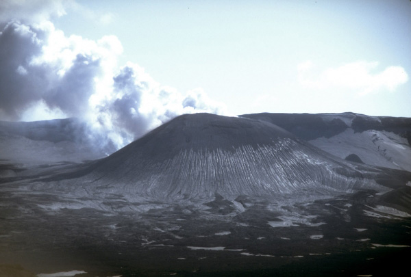 View of the steaming cinder cone that marks the site of the most recent eruptive activity at Okmok caldera, a 9.3-km (5.8 mi)-diameter circular crater that truncates the top of a large shield volcano on the northeastern part of Umnak Island in the eastern Aleutian Islands. Eruptions from this cone in 1945 and 1958 produced lava flows that extruded onto the caldera floor. Photograph by C. Nye, Alaska Division of Geological and Geophysical Surveys, September, 1980.