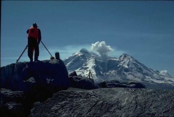 Geologist John Ewert uses a theodolite to measure the dimensions of the lava dome at Redoubt Volcano.  From the same installation, scientists also utilized a laser-ranging device to measure distances to reflectors.  Repeated measurements of these distances allow for detection of ground deformation that may be related to volcanic activity.