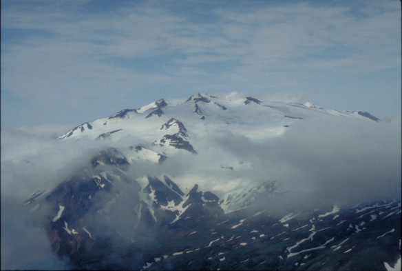 Broad summit of Makushin Volcano as seen from the north side of Unalaska Island.  Smoke from the summit fumaroles is faintly just left of center of summit.  The sharp ridges to the right are the result of Pleistocen to Holocene glaciation of the volcano massif.  Broad grassy foreground are Holocene andesite that reach the Bering Sea.