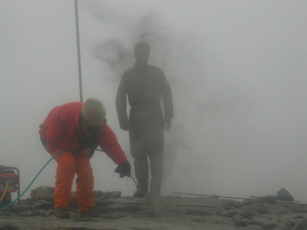 Creating station OKSO (Okmok South).  "Max in the mist" -- Nicole and Max cleaning out the drill hole during the installation of a permanent GPS station at OKSO.
