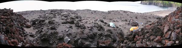 Panoramic photo of the volcaniclastic debris flow fan deposit that was formed by the 2001 eruption. The panoramic covers the view from the west to the northwest. The photo was taken from about half way up the central part of the fork in the northern portion of the largest 2001 lava flow on western flank of Mt. Cleveland. The a'a lava flow can be seen on the left and right edges of the photo. The location of the 2003 field camp is visible in the photo right of center. The larger breadcrust bombs that make up part of the newly formed volcaniclastic debris flow fan deposit are observed in the foreground and extend straight out to the west. The height of the breadcrust bombs in this area are likely the cause for the forking of the lava flow.