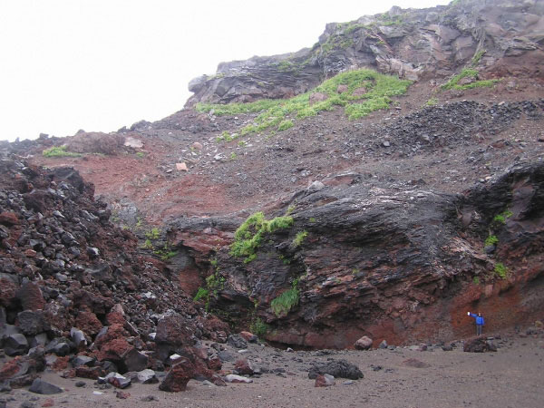 Photo of a stratigraphic section located just south of the southern secondary forking lobe of the 2001 lava flow (visible on the left side of the photo) that was deposited on top of the newly formed volcaniclastic debris flow fan deposit from the 2001 eruption. Steve J. Smith for scale in the lower right of the photo (1.8 meters tall). The section displays at least two previous depositional episodes in the volcano's past of producing (from bottom to top) clast supported debris, basal breccia, and lava flow. This is similar to the depositional sequence of the 2001 eruption on the western flank.