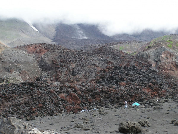 Photo taken from the northwestern part of the newly formed volcaniclastic debris flow fan deposit from the 2001 eruption looking toward the southeast at breadcrust bombs. The northern fork of the main 2001 lava flow is observed in the background with the gray and teal colored tent (2-man Mountain Hardware Trango for scale) to the lower right of center indicating the location of the camp. The location of the camp is where this northern fork reached the highest part of the fan deposit, and forked for a second time. On the upper portions of the lava flow there is some steaming observed almost in the channel of the lava flow. This is where some fumarolic activity is taking place from the seeping of rainwater into the cracks of the flow.