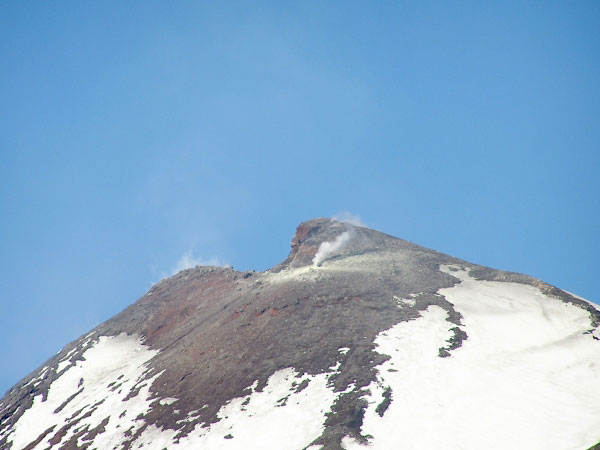 A zoomed in view from camp of the east-northeastern summit region of Mount Cleveland. Fumarolic steam is observed from the southern part of the crater rim (left side of the photo), and from a vigorous fumarole on the eastern side of the summit with some sulfurous deposits also observed (a little downslope from the crater). This view shows the collapsed region of the southern portion of the crater rim (left side of the summit). There was no cut out portion observed by the field party in September 2002. The cut out (collapsed) part was first observed from a similar vantage point - though further out to sea - but due to more cloud cover that day, it was difficult to be certain of the change. This view in 2004 confirmed that some morphologic change took place at the summit since 2002. This photo was enhanced to bring out better contrast.
