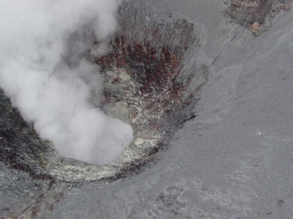 Aerial view into the summit crater of Korovin. This level of fumarolic emission from the slightly oblong, 60&ndash;90 m (200&ndash;300 ft) diameter, sheer-walled orifice at the bottom of the crater is apparently fairly typical and can account for the periodic observations of plumes from Atka Village. At times, a shallow body of gray, turbid water partially fills the inner crater and, in 2004, was observed roiling. Phreatic explosions from this water-rich, high-temperature system may be responsible for the occasional localized ash-fall deposits seen on the upper flanks of Korovin. Photograph by R.G. McGimsey, AVO/USGS, July 19, 2004.