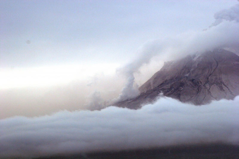 Augustine Volcano on 1/29/06 observation overflight.  Photo from the west with a view of the north and west flanks of the volcano. White steam and gas on the north flank is from a recent pyroclastic flow deposit on the north flank (probably emplaced within an hour of the photo being shot).  The main ash and gas plume is drifting to the S SW.
