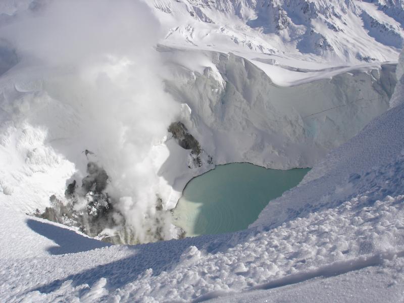 View from the summit ridge of Mt. Spurr down into the ice cauldron.  Part of the turquoise lake, bare warm steaming ground (rock?), and steam rising off of fumaroles are visible.  Note that multiple point-source fumaroles are venting on the prograding(?) delta along north (left) side of lake.