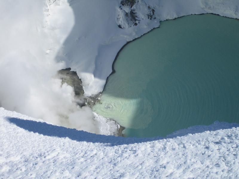 View from the summit ridge of Mt. Spurr down into the ice cauldron.  The turquoise lake, bare warm steaming ground (rock?), and steam rising off of fumaroles are visible.  Note also the roiling spring adjacent to the fumaroles and the change in water color emanating from it.    