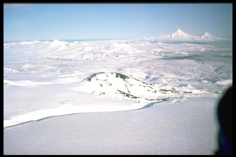 Aerial view northeast from a position near Westdahl Peak of the snow-filled fissure and cinder cone produced by the 1991 eruption of Westdahl volcano.  The moderate peaks in the middle distance are Fisher Caldera.  The two prominent peaks in the upper right are Shishaldin (left) and Isanotski (right).
