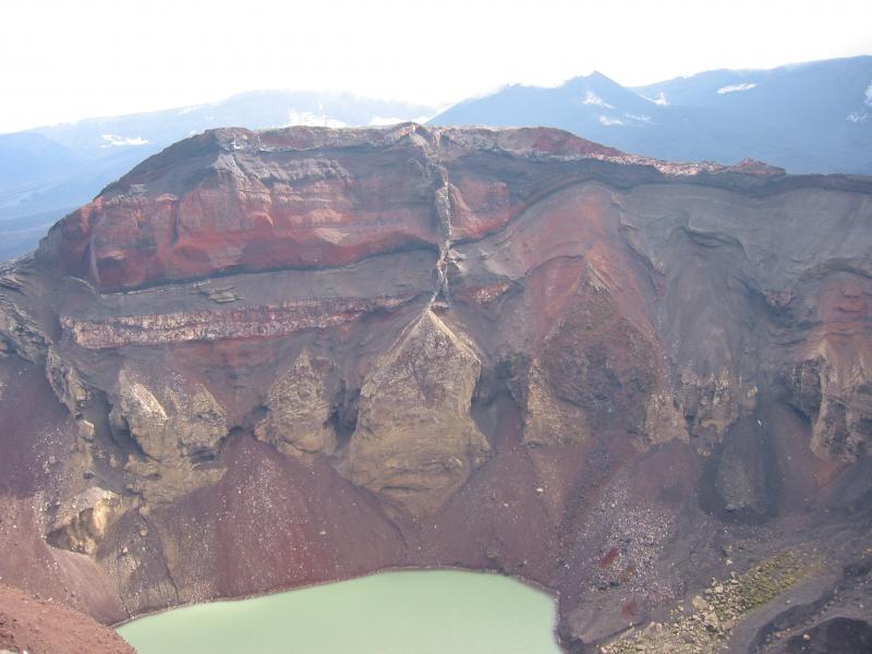 View of the south inner wall of Cone E, a post-caldera vent structure at Okmok Caldera in the eastern Aleutians.  Cone E formed during multiple eruptions that produced tephra and lava flows; it collapsed in a violent explosive eruption several hundred years ago to form this crater that now contains a small lake about 425 x 200 m across.  The far wall exposes the insides of Cone E and consists of layers of lava, spatter, tephra, and cross-cutting feeder dikes, or frozen conduits for magma&rsquo;s ascent to the surface.  