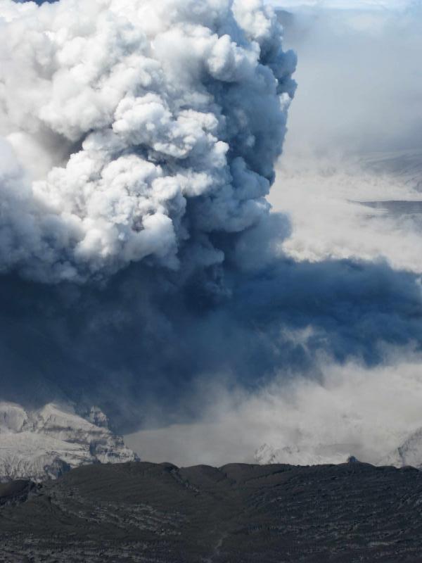 A look down into the caldera from above the east rim of Okmok.  The photo shows a tan-colored lake and white steam plumes and gray ash plumes eminating from vents near the base of cone D.
