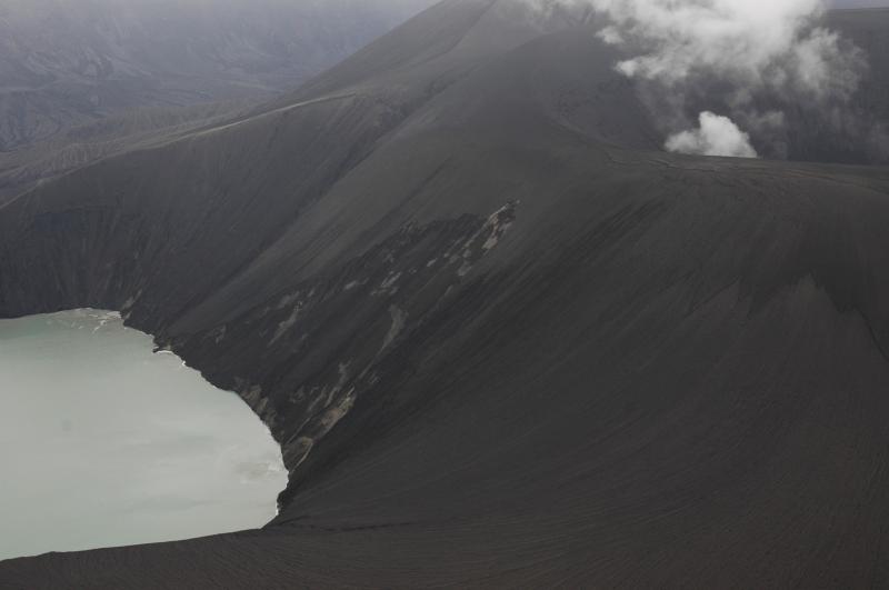 view to the southeast across the modified old lake and the northeast flank of the steaming new tephra cone built atop the Cone D lava bench.  We estimate that the cone rim shown here is about 800 feet in elevation above the lake surface.  Lighter colored material exposed in the slumped face is dry pyroclastic material.  One of at least two zones of fumarolic upwelling is visible in the far bay of the lake.  Above this zone, note the dark layers of Cone D lavas newly exposed, we think, by explosions during the 2008 eruption. 