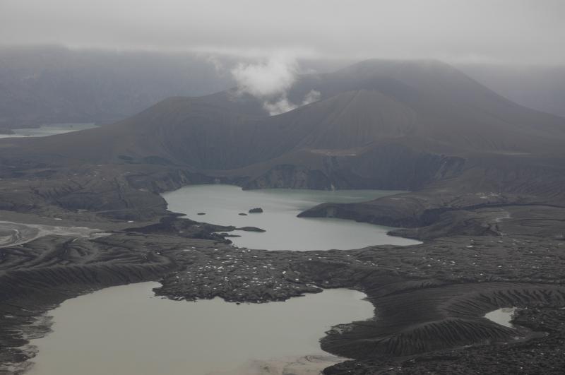 Foggy day inside Okmok looking across the Cone G lake, the speckled ponds on the surface of the 1997 lava flow, and the new explosion crater lake at the west base of the new tephra cone complex. The larger tephra cone emits a puffing plume of steam and minor amounts of volcanic gas. Cone D tickles the cloud deck beyond and the modified old lake is visible at the upper left.  