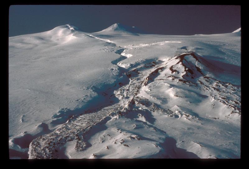 Westdahl Peak, in upper left corner of image (Faris Peak in distance), was the starting point of the fissure (now appearing as sinuous ice crevasse) that extends to the cinder cone (middle right),  produced in the 1991 eruption.  Lava flows were fed from drainage of the fissure and from the newly formed cinder cone. 