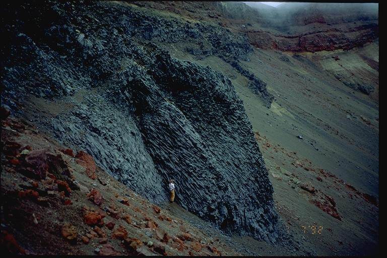 					Aniakchak 1992. Keri Petersen stands below the jointed columnar lava flows that compose the west wall of Half Cone. 			Photograph courtesy of Game McGimsey. 