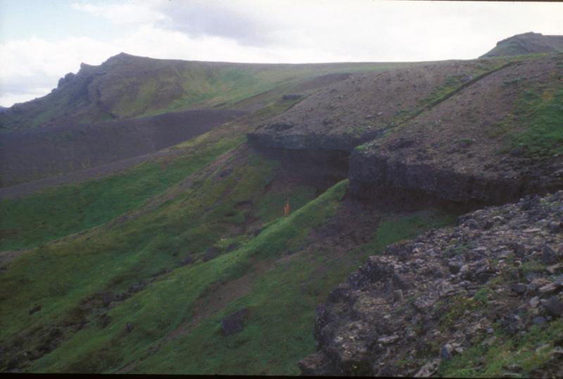 Deposits from the caldera-forming eruption 9,400 years ago, about 5 kilometers outside the eastern caldera wall.  AVO scientist Dr. Jim Gardner stands near the base of ~6.5 meters of pumice fall, capped by ~2.5 m of densely welded pyroclastic flow material.  Additional pyroclastic flow material above is obscured by slope wash.  In the foreground is the top of the welded portion of pyroclastic flow deposit.