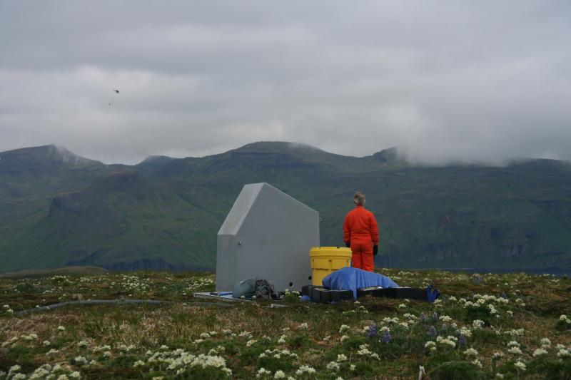 AVO field technician Ed Clark waits for the next sling load of equipment to be delivered to seismic station AKSD on Akutan. Ed's orange flightsuit is made of flame-resistant Nomex: essential gear for helicopter operations.