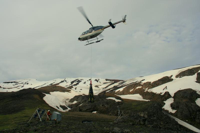 Upgrading the radio and power systems at remote Akutan Volcano via helicopter sling load. Loads carry batteries, solar panels, and concrete to each installation site. In this photograph, AVO field technician Cyrus Read watches as a sling load of batteries is delivered to station AKLV.