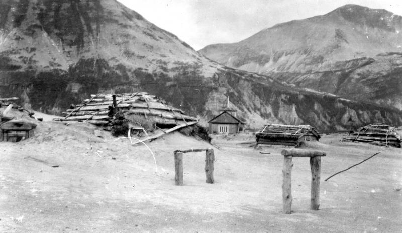 Photograph of ash drifts around Katmai village barabaras (sod houses) after the June 1912 eruption of Novarupta. Katmai's then-new Russian Orthodox church is visible in the background. Photo taken by G.C. Martin, U.S. Geological Survey. This image is image mgc00747 in the U.S.G.S. photolibrary.