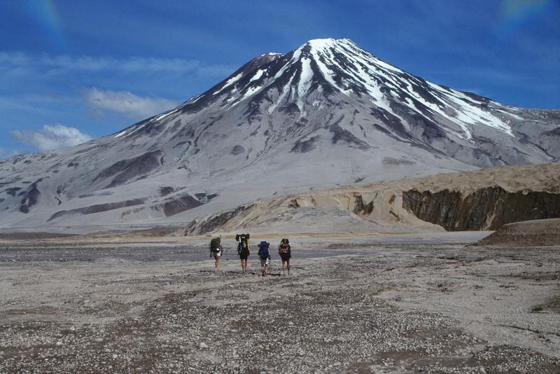 Geologists walk toward 2,317-m (7,602 ft)-high, Mount Griggs volcano from the base of Knife Creek Glacier at the base of the northwest flank of Mount Katmai. Mount Griggs volcano lies 10 km (6 mi) behind the volcanic arc defined by other Katmai group volcanoes.
Although no historical eruptions have been reported from Mount Griggs, vigorously active
fumaroles persist in a summit crater and along the upper southwest flank. The slopes of Mount Griggs are heavily mantled by gray fallout from the 1912 eruption of Novarupta volcano. View is to the north. Photograph by G. McGimsey, U.S. Geological Survey, July 16, 1990.  L-R, Susan Walker, John Paskievitch, John Eichelberger, Jack Kleinman.