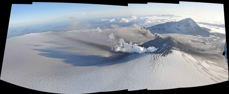 Aerial view toward the southwest of the actively erupting cone within Veniaminof caldera.  The white steam plume is produced where a lava flow is descending the side of the cone and melting snow and ice.  The darker colored, ashy plume is rising in bursts from the active vent. Nearly continuous eruption from this vent since early June has built a new small tephra cone and an apron of lavas along the southwest flank of the cone. The caldera is about 10 km (6.5 mi) in diameter.  The cone rises about 330 m (1080 ft) above the surrounding ice field.  Note the dark stripe of ash fall on the ice.  This photo was taken during a brief helicopter trip into the caldera by AVO Geologist Game McGimsey and Dickinson College Geology Professor Ben Edwards.  