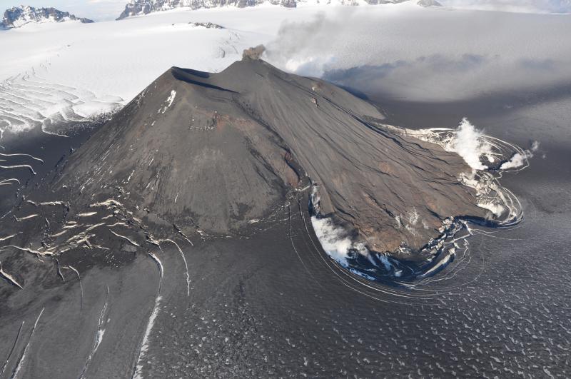 A small puff of ash emerges from the active cone inside Veniaminof caldera on August 18, 2013.  A beautiful fan of lava flows active earlier in the summer descends the south flank of the cone onto glacial ice producing white steam clouds and depressions where melting has occurred.  The surrounding glacier is darkened by recent ash fall.  