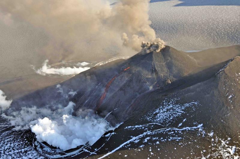Aerial view of the eruption at Veniaminof&#039;s intracaldera cone, August 18, 2013. This cone rises about 1000 feet above the surrounding icefield.  It has been intermittently erupting lava, ash and steam since June 13, 2013. This photo shows the incandescent, orange stream of molten lava emerging from the active cone.  Steam billows from the pit at the base of the cone where the lava encounters and melts ice and snow. A small, ash-rich plume rises just above the vent producing a diffuse ash cloud that drifts downwind.  In the foreground, round white patches probably represent ballistic impact craters. Photo taken by Game McGimsey, AVO/USGS. This overflight of Veniaminof was co-sponsored by the National Geographic Society.