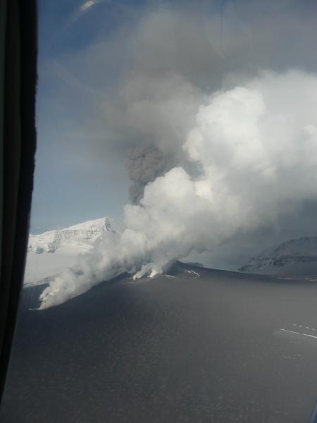 Aerial view of Veniaminof volcano in eruption at about 11:30 am on September 7, 2013.  Photograph by Joyce Alto.  White water vapor clouds rise from regions where hot lava is encountering snow and ice.  A gray-brown ash column rises in the background from the active vent. Note the ash-darkened icefield within the summit caldera.