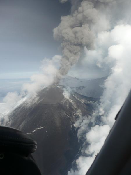 Aerial view of Veniaminof volcano in eruption on September 7, 2013. Photograph by Joyce Alto.  The gray brown ash laden eruption column ascends from the active vent at the top of the cone.  White vapor clouds are formed where lava is melting snow and ice.  
