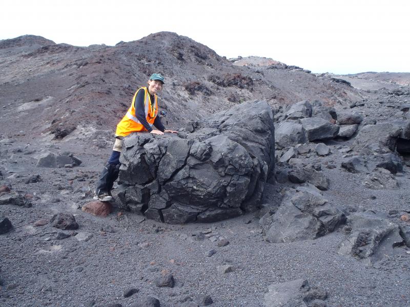 Kirsten Nicolaysen, Whitman College and Islands of Four Mountains Project leader, leans on a recently emplaced lava block carried down the flank of Cleveland volcano in a block and ash flow.  The deposit probably formed when a lava flow high on the flank of the volcano collapsed and tumbled down the side of the volcano, all the way to the sea. Photo taken during the 2014 field season of the Islands of Four Mountains multidisciplinary project, work funded by the National Science Foundation, the USGS/AVO, and the Keck Geology Consortium.