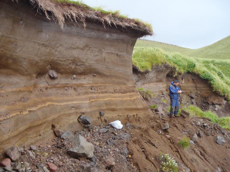 Mitsuru Okuno stands next to a sequence of young tephra falls atop coarse lahar deposits on the southeast coast of Carlisle Volcano.  Photo taken during the 2014 field season of the Islands of Four Mountains multidisciplinary project, work funded by the National Science Foundation, the USGS/AVO, and the Keck Geology Consortium.