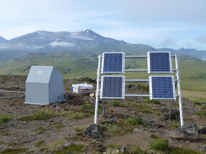 Seismic station CLCO, Concord Point, Chuginadak Island. The peaks of  Tanax̂ Angunax̂ volcano are in the background. The solar panels on the swingset frame in foreground are part of the power system for CLCO and a webcam out of sight to the left. Photograph from fieldwork in the Islands of Four Mountains, John Lyons, USGS/AVO, August 1, 2014.
