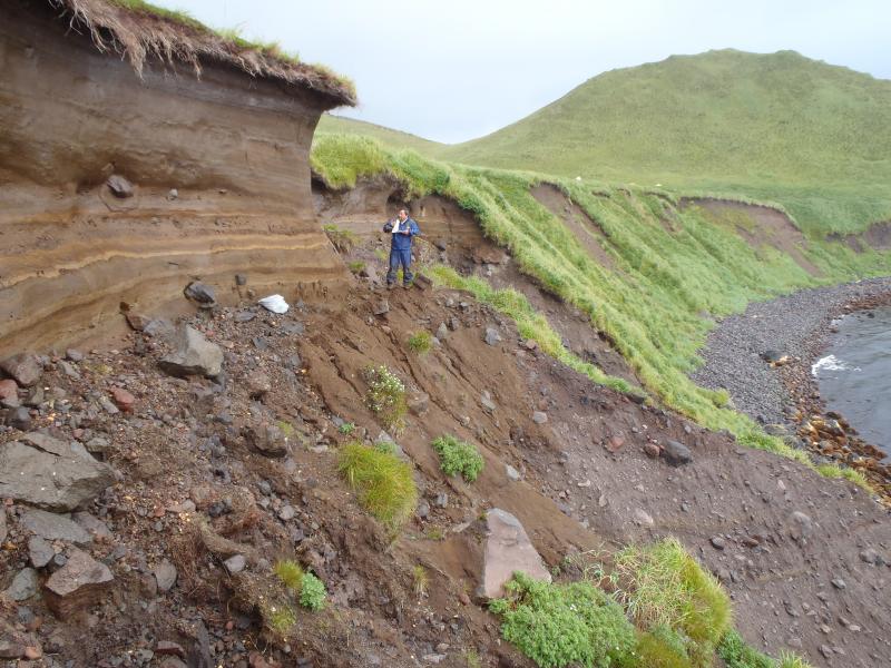 Mitsuru Okuno and tephra section in coastal bluff, east side of Carlisle Island. Photo taken during the 2014 field season of the Islands of Four Mountains multidisciplinary project, work funded by the National Science Foundation, the USGS/AVO, and the Keck Geology Consortium.