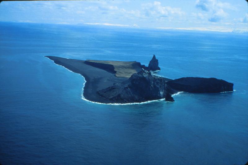 Aerial view, looking south, of Bogoslof Island, which is the summit of a largely submarine stratovolcano located in the Bering Sea 50 km (31 mi) behind the main Aleutian volcanic arc. The island is about 1.5x 0.6 km (1 x 0.4 mi) and, due to energetic wave action and frequent eruptive activity, it has changed shape dramatically since first mapped in the late 1700&#039;s. Its most recent eruption, in 1992, produced the conical, rubbly lava dome (150 m [492 ft] high)and offshore spire at bottom center. Photograph by T. Keith, U.S. Geological Survey, May 10, 1994.