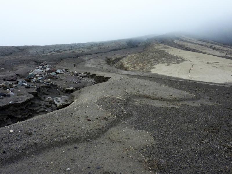 Deposits at the mouth of Peregrine ravine on Kasatochi.  The lightest-colored deposits, traversed by footsteps, are undisturbed 2008 eruption deposits.  The mottled darker deposits immediately to the left of the footsteps are deposits of debris flows composed of the uppermost, fine-grained portion of 2008 deposits that formed in the waning stages of, or immediately after the eruption.  The foreground and left side of the image show overlapping deposits of post-eruption debris flows composed mostly of water-mobilized 2008 deposits which accumulated in the ravine and were episodically transported downstream. See http://doi.org/10.14509/29718 for additional details.