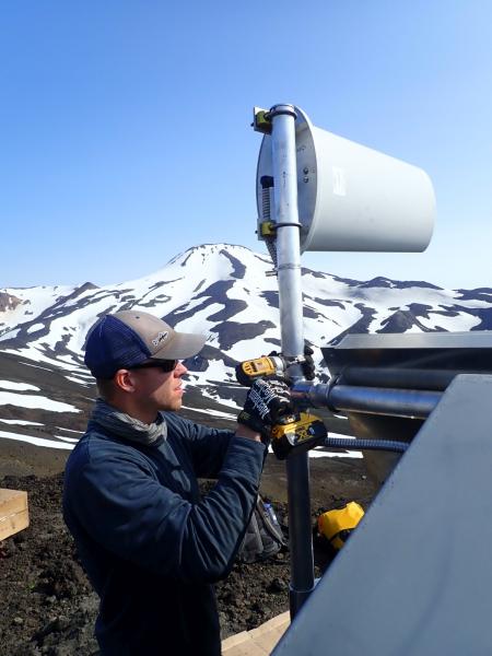 John Lyons (AVO-USGS) secures an antenna cable at the upgraded seismic station KOKV on the south flank of Korovin Volcano, Atka Island.  The snowy NW side of Kliuchef volcano fills the background.