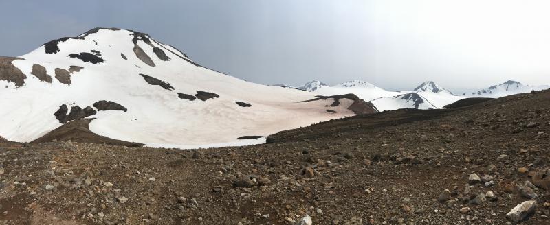 The circular depression in the center of view, on the high southern flank of Kliuchef volcano (high peak to the left) is interpreted as a previously unmapped vent; the feature was encountered by Janet Schaefer (AVO/ADGGS) and Michelle Coombs (AVO/USGS) during the 2019 field mapping project of the Atka volcanic complex.  Photo taken from station 19AAJRS009.  A sample from spatter lava blocks was taken from the WNW rim of the crater at station 19AAJRS008, visible in this photo on the left rim of the depression.