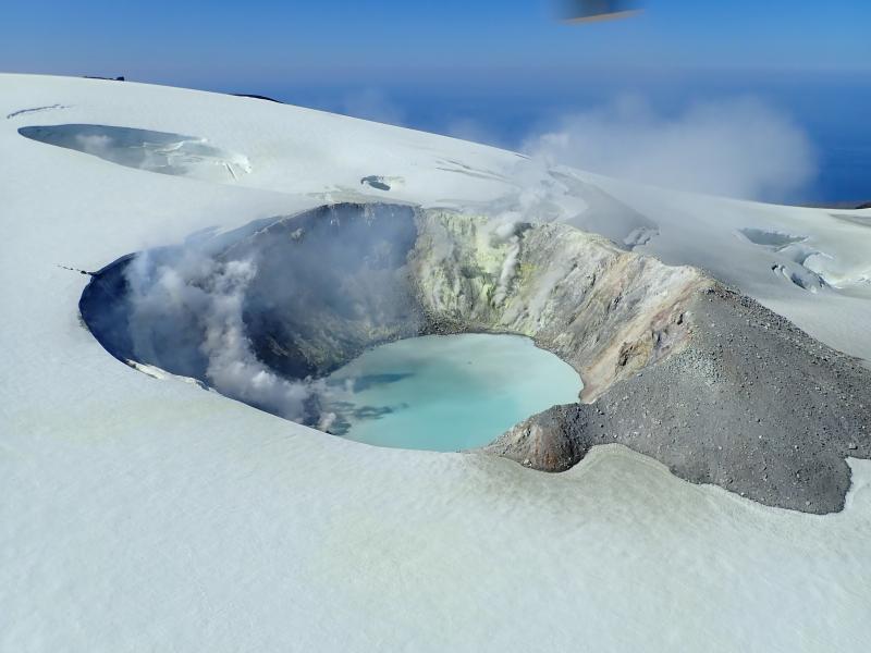 August 2019 views of the summit crater of Makushin Volcano.  Heat from vigorous fumarolic activity is melting depressions,  holes,  and caves in the glacial ice.  A low plume of sulfurous volcanic gases drifts off to the NNE.