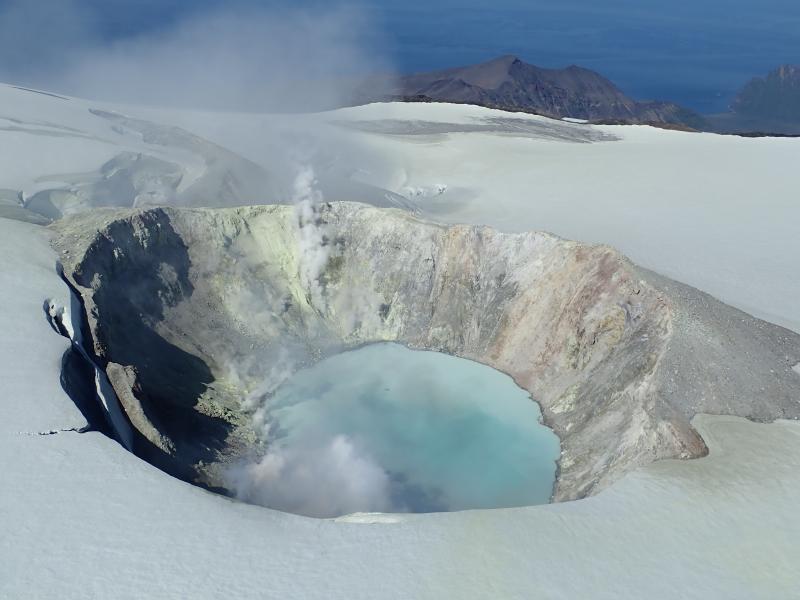 August 2019 views of the summit crater of Makushin Volcano.  Heat from vigorous fumarolic activity is melting depressions,  holes,  and caves in the glacial ice.  Sulfur rich mineral precipitates paint the ground yellow near fumaroles.  Pastel earthen tones  of altered rock and blue lake water add to the palette of colors.