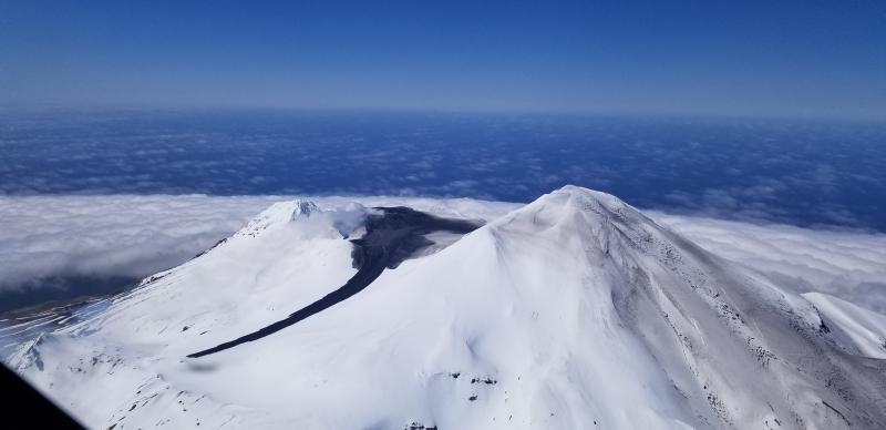 Summit crater of Great Sitkin volcano, May 30, 2021. Dark linear feature in middle foreground is a lahar (volcanic mudflow) deposit. Note trace ash fall over snow on right flank of Great Sitkin summit. The dark deposits in the summit crater are likely ash fall and pyroclastic flow deposits.