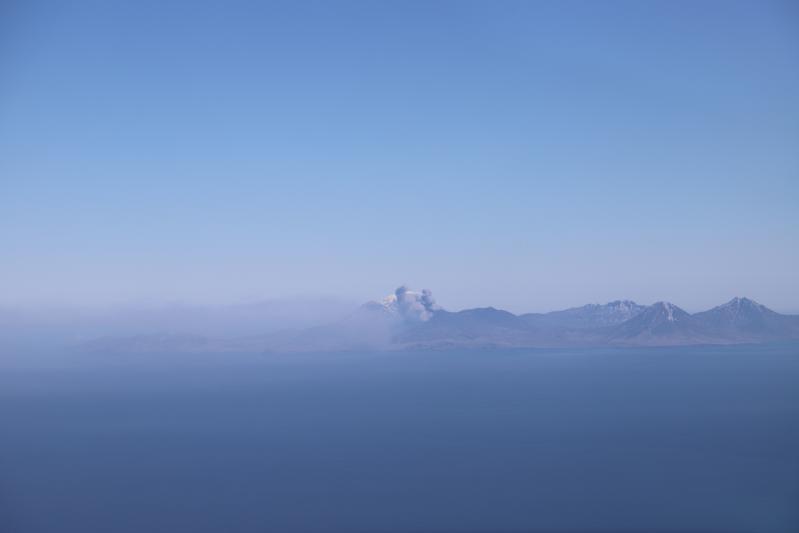 The ash plume from the eruption from the North Crater of Mount Cerberus at Semisopochnoi on May 30, 2021 as viewed from the southeast in a helicopter. The diffuse ash plume extends across the image as a gray haze (to the southwest). Two distinct puffs to a higher altitude are visible near the vent.