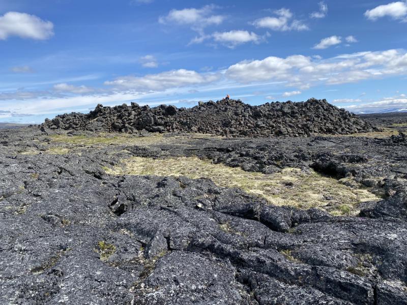 Ground view of a shatter ring on the Lost Jim lava flow, with seated geologist for scale. Shatter rings form over active lava tubes due to fluctuations in discharge, which causes the lava tube roof to flex repeatedly up and down.