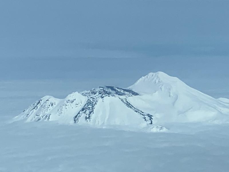Photograph of Great Sitkin Volcano, showing lava flows that have been forming during the eruption that started in May 2021. Recent lava effusion has formed the relatively snow-free lava lobes covering the right (eastern) side of the summit crater. Photo by Dave Clum, Alaska Airlines pilot, May 20, 2023. 