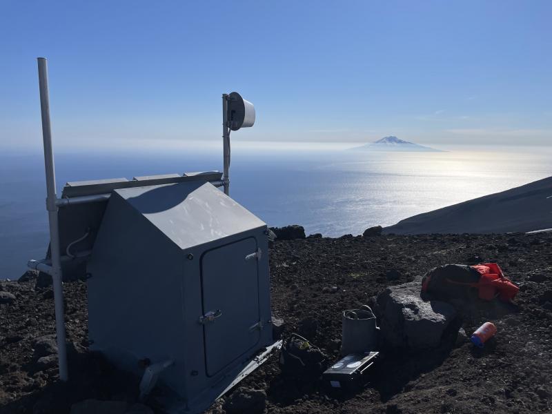 AVO field crew making repairs at site TASO on the south flank of Tanaga Volcano, on a rare sunny evening.  Gareloi Volcano is visible in the distance.