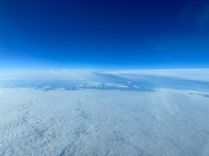 Photo of Shishaldin Volcano (center-right) on July 18, 2023, as an ash-rich plume erupted, taken from the air. View from the north of Shishaldin, with the ash-rich plume extending to the southeast. Isanotski volcano (center) and Roundtop (center-left) are also visible.