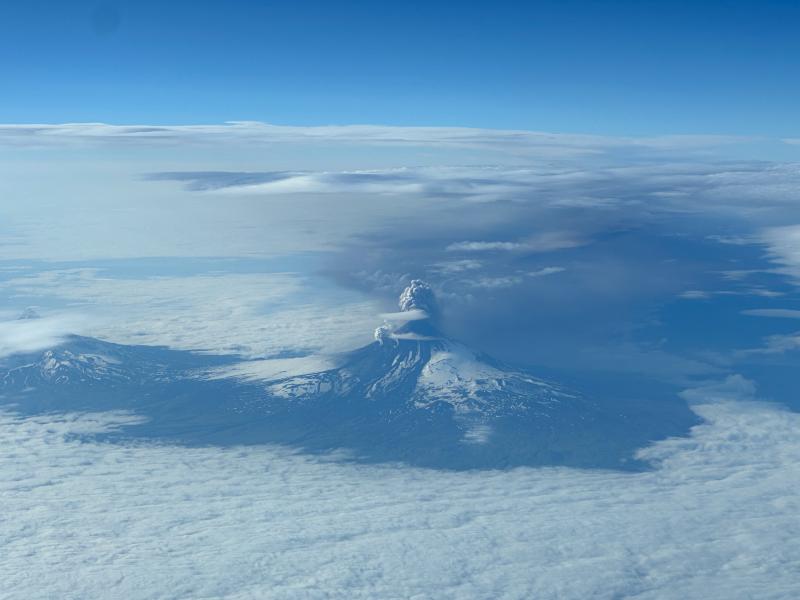 Photo of Shishaldin Volcano on July 18, 2023, as an ash-rich plume erupted, taken from the air. View from the north of Shishaldin, with the ash-rich plume extending to the southeast. Isanotski volcano is visible on the left-hand side of the image, partially obscured by clouds.